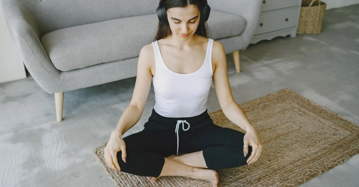 Femme méditant à l'intérieur sur un tapis avec des écouteurs, favorisant la détente et le bien-être.