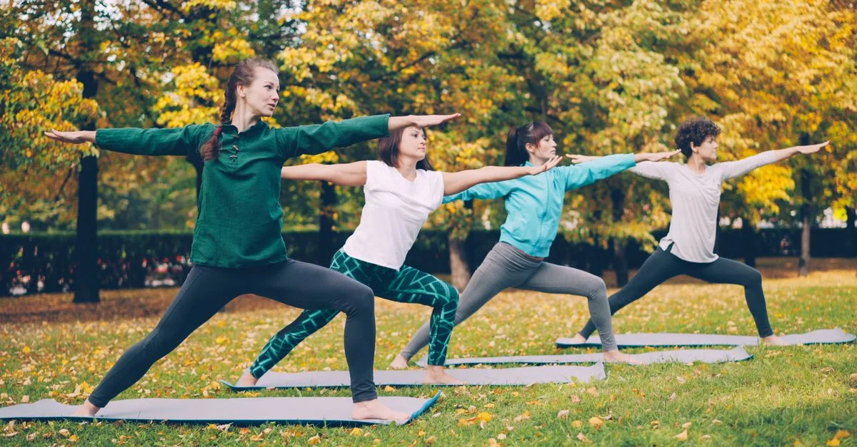 Quatre femmes pratiquant le yoga dans un parc entouré de feuillage automnal.