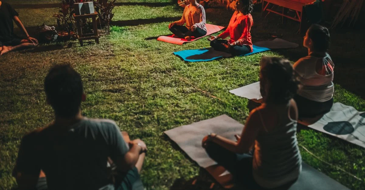 Un groupe d'adultes pratiquant le yoga en plein air la nuit sur des tapis dans un cadre de jardin serein.